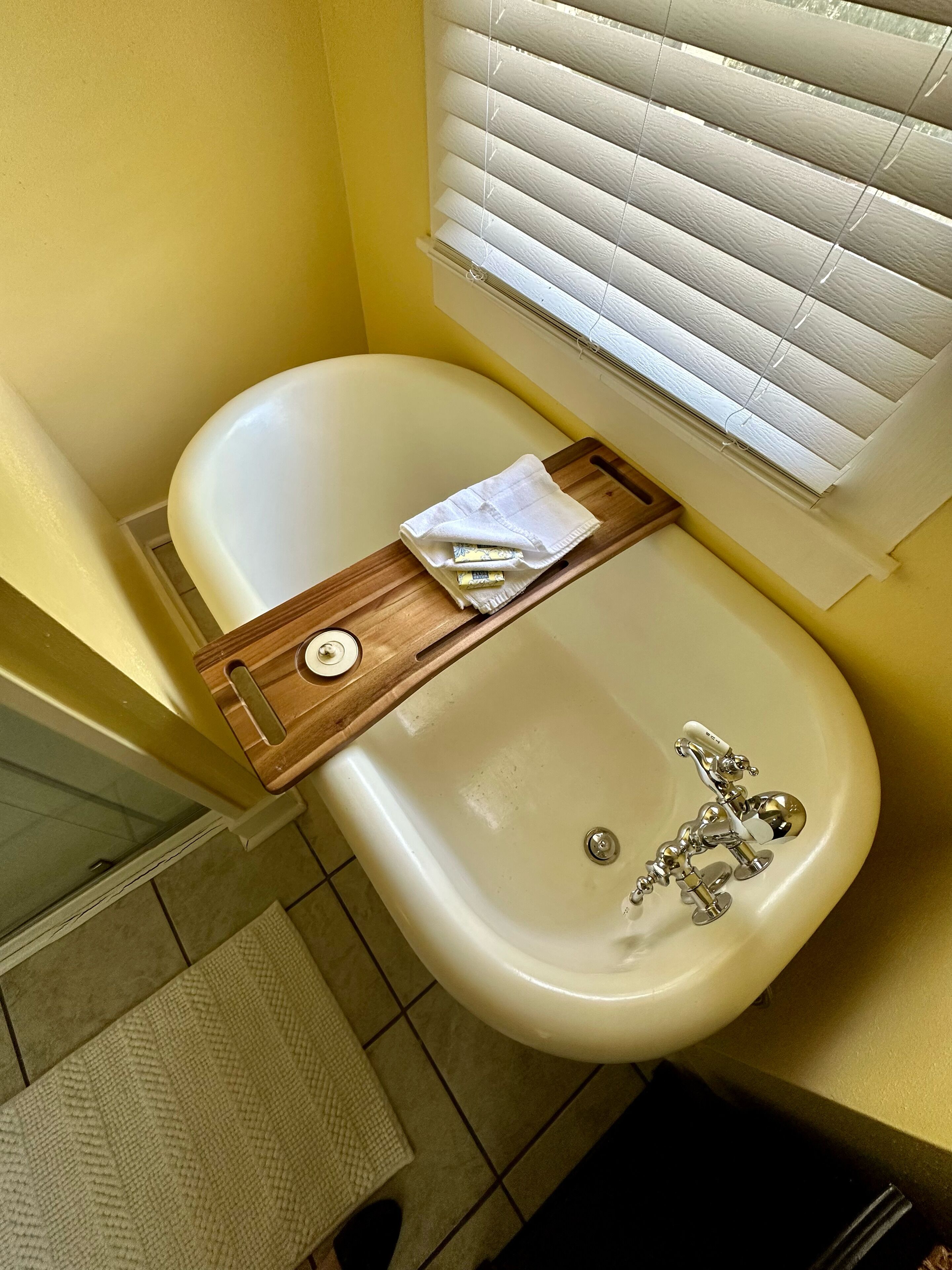 Une salle de bain avec une baignoire blanche, un plateau en bois contenant des articles de toilette et une fenêtre avec des stores.