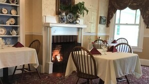 Ein Esszimmer mit zwei Tischen, Stühlen, einem Kamin und einem Bücherregal.
