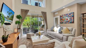 A spacious living room with a sofa, coffee table, and a TV on a stand. There is a balcony with a view of greenery and a pool. - Magic Village Resort 4 Bedroom townhome with Outdoor Kitchen (Kissimmee)