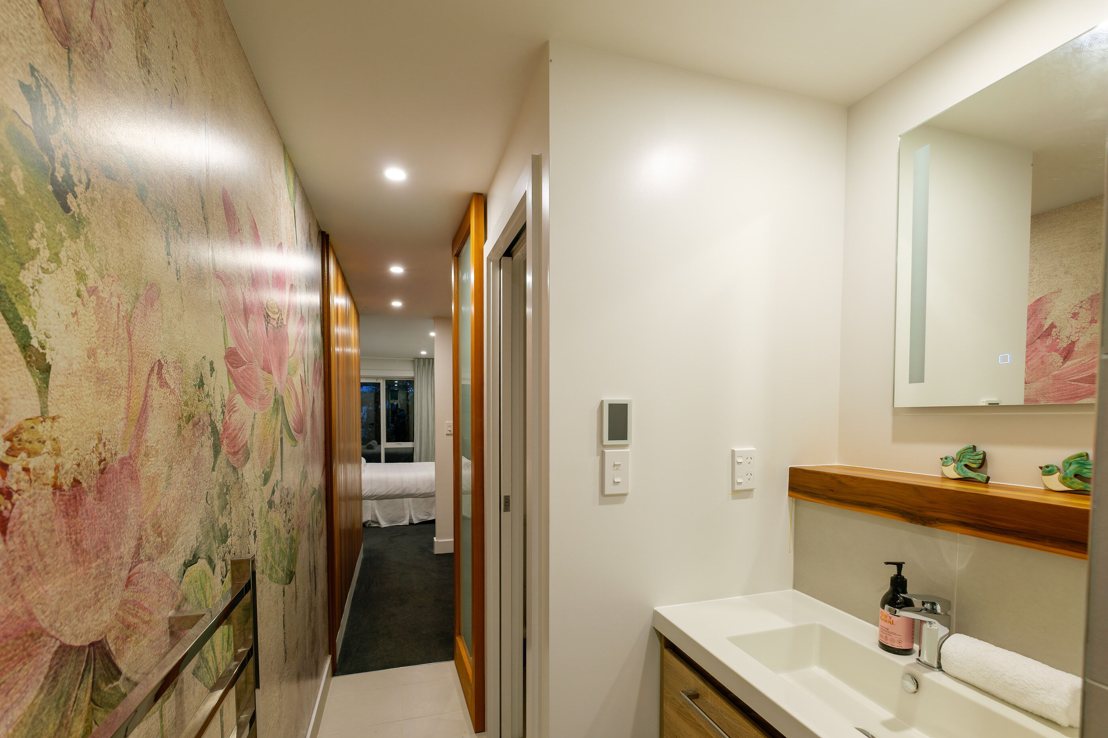 Un pasillo de hotel con un mural floral, un baño con lavamanos y espejo blancos, y una cama al fondo.