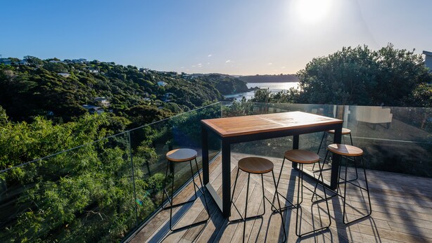 A wooden deck with a table and bar stools overlooking a scenic view of a lake and hills. - The Kingfisher House - Oneroa (Waiheke Island)
