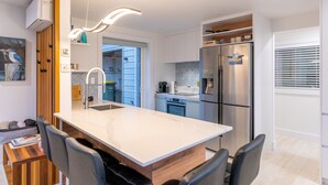 A modern kitchen with a central island, stainless steel appliances, and a dining area with chairs. - The Kingfisher House - Oneroa (Waiheke Island)