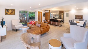 A spacious living area with a sofa, chairs, a dining table, and a kitchen in the background. - The Kingfisher House - Oneroa (Waiheke Island)