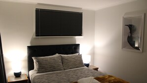 A neatly made bed with a headboard, two bedside lamps, a window with blinds, and a framed artwork on the wall. - Orchard Leigh on the Quarry (Dunedin)