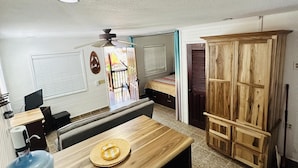Habitación pequeña con una mesa de madera, dispensador de agua, ventilador de techo, cama con ventana y armario.