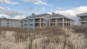 Ein Hotel direkt am Strand mit mehrstöckigen Gebäuden, Balkonen und einem sandigen Vordergrund mit vereinzelter Vegetation.
