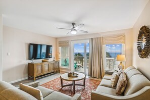 Sala de estar com televisão de tela plana, ventilador de teto, sofÔ, mesa de centro e vista para o exterior.