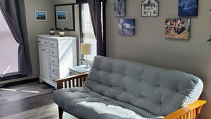 Ein modernes Zimmer mit grauem Sofa, einer hölzernen Bank und einem weißen Schminktisch. An den Wänden hängen Bilderrahmen und es gibt ein Fenster mit Vorhängen.