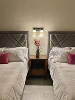 A hotel room with two beds, a nightstand with a vase of flowers, and a wall-mounted light fixture.