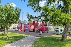 A playground with red and blue play structures, a covered seating area, and a paved path.