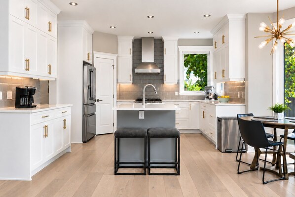 A modern kitchen with white cabinetry, a stainless steel refrigerator, a built-in oven, a central island with a sink, and a dining area with a table and chairs. - Luxury Estate - Stampede & Summer Oasis (Calgary)