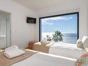 A hotel room with a large window, a flat-screen TV, two beds, a small table, and a view of the sea.