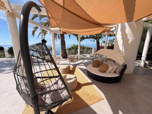 A covered outdoor seating area with a swing chair, cushioned seating, and a view of the sea.