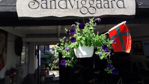 Ein hölzernes Schild mit der Aufschrift 'Sandvigaard' und ein Blumentopf mit violetten Blüten.