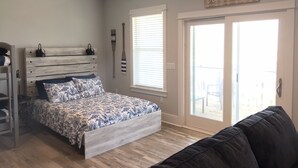 Ein Schlafzimmer mit einem Bett, einer Kommode, einem Deckenventilator, einem Fenster mit Jalousien und einer Glasschiebetür, die zu einem Außenbereich führt.