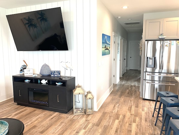 A modern kitchen with a stainless steel refrigerator, wooden flooring, and a flat-screen TV mounted on the wall.
