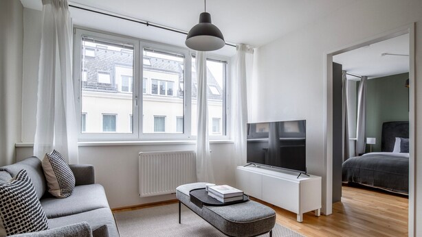 A modern living room with a sofa, a coffee table, a TV, and a view of the city through large windows. - Premium Hernals 1BR w/ Balcony, nr Lidlpark, by Blueground (Vienna)