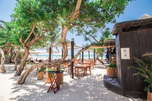 A beachside restaurant with outdoor seating, wooden tables, and chairs, surrounded by trees and overlooking the ocean. - HAU Holbox Beach Front (Isla Holbox)