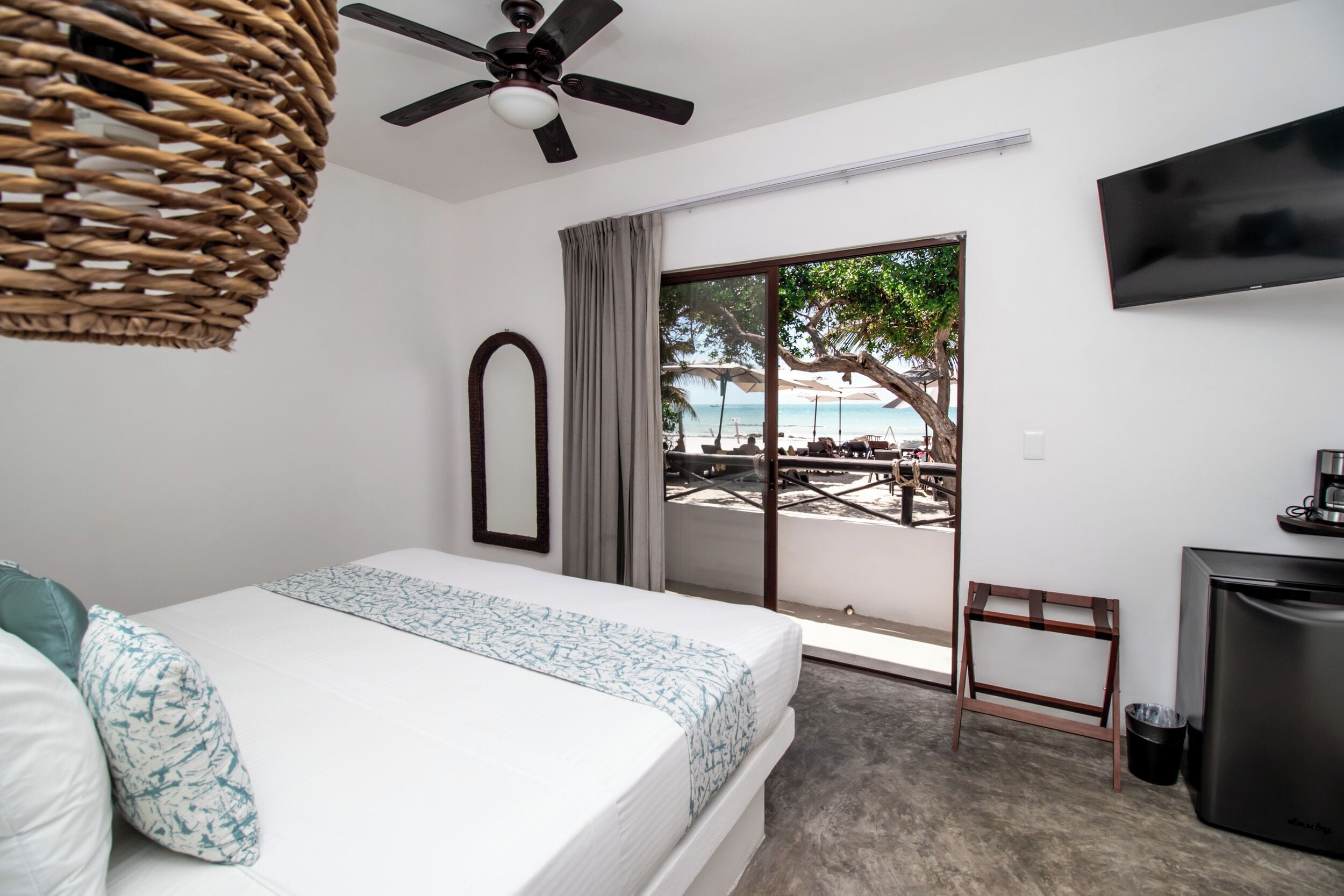 A hotel room with a bed, a ceiling fan, a TV, a small desk, and a view of the beach through the window.