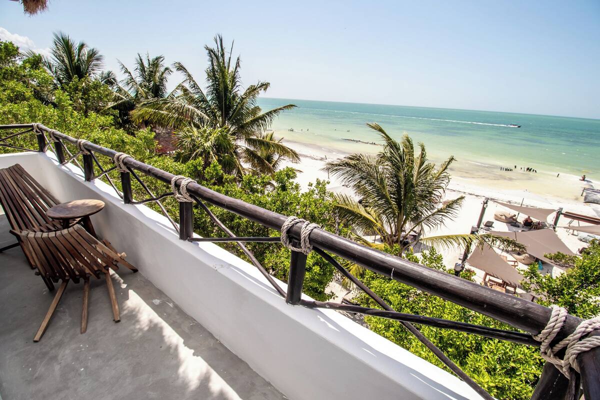 Un balcón con una silla y mesa de madera, con vista a una playa con palmeras y cielo despejado.