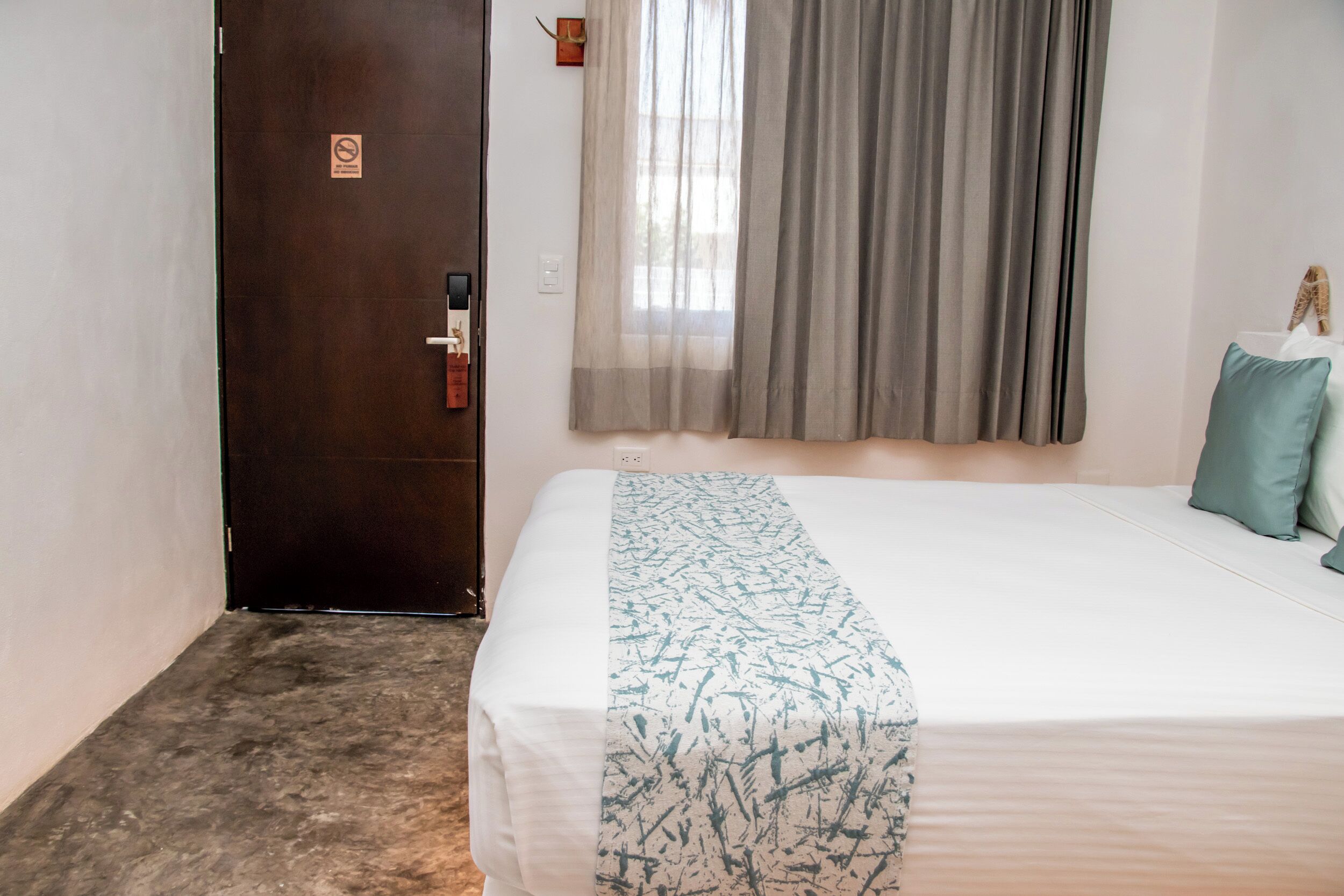 A hotel room with a bed, a door, a window with curtains, and a wall-mounted light switch.