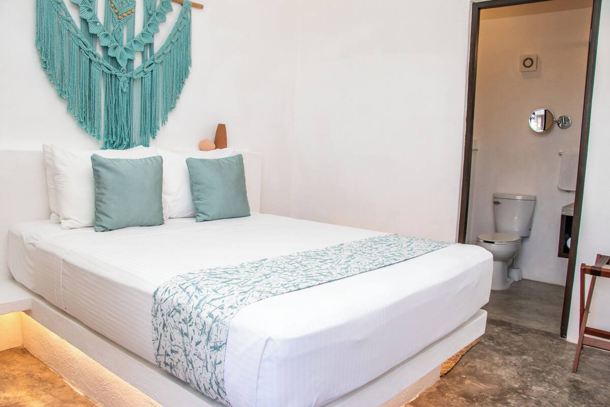 Un dormitorio con una cama, un colgador de macramé en la pared y un inodoro visible en la habitación contigua.