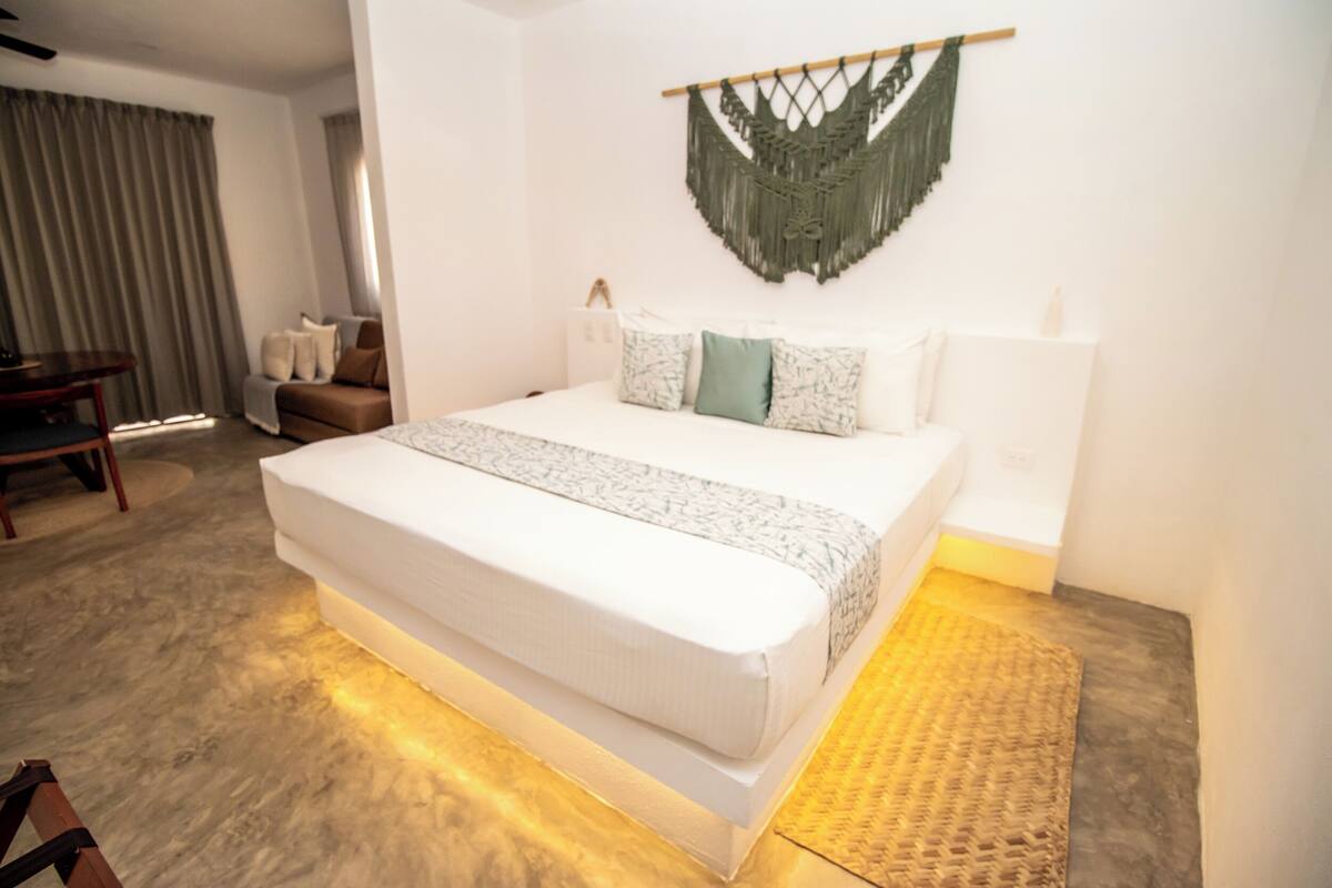 Una habitación de hotel moderna con una cama grande, suelo de madera, un tapiz de macramé y vista al interior de la habitación.