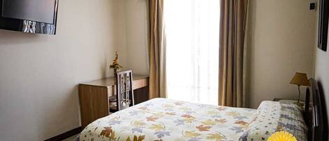 A bedroom with a bed, a desk, a chair, a TV mounted on the wall, and a window with curtains.