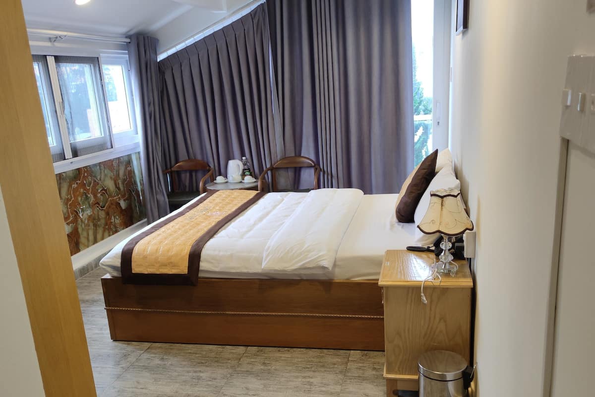 A modern hotel room with a large bed, a wooden nightstand, a desk, a chair, and a view of the outdoors.