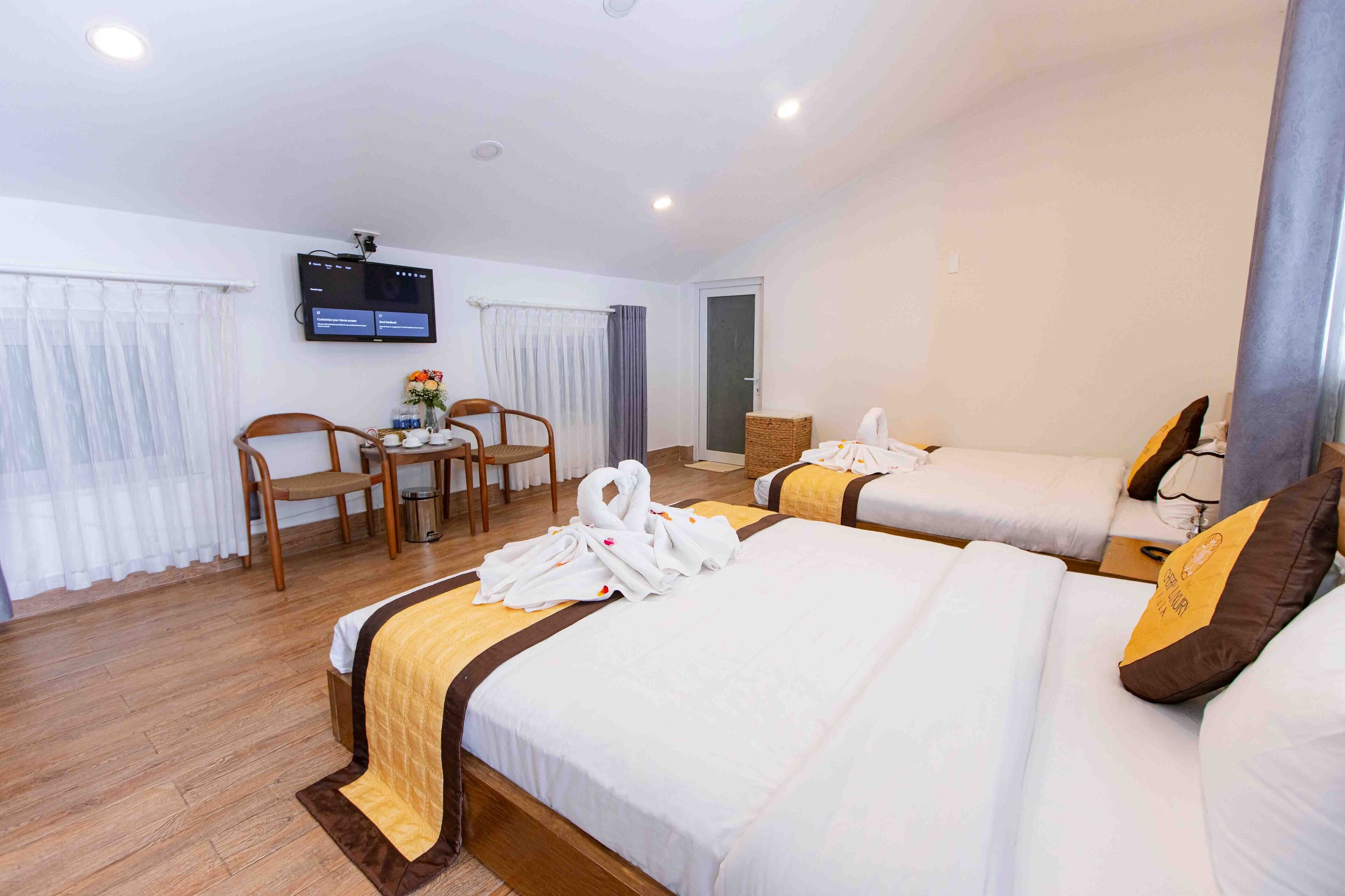 A hotel room with two beds, a TV, a desk with a chair, and a small table with a vase of flowers.