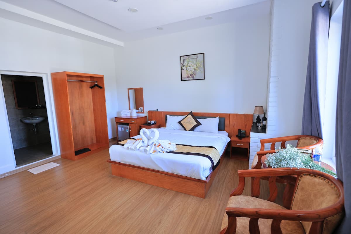 A hotel room with a wooden bed, a wooden chair, a desk with a lamp, and a small table with a plant.