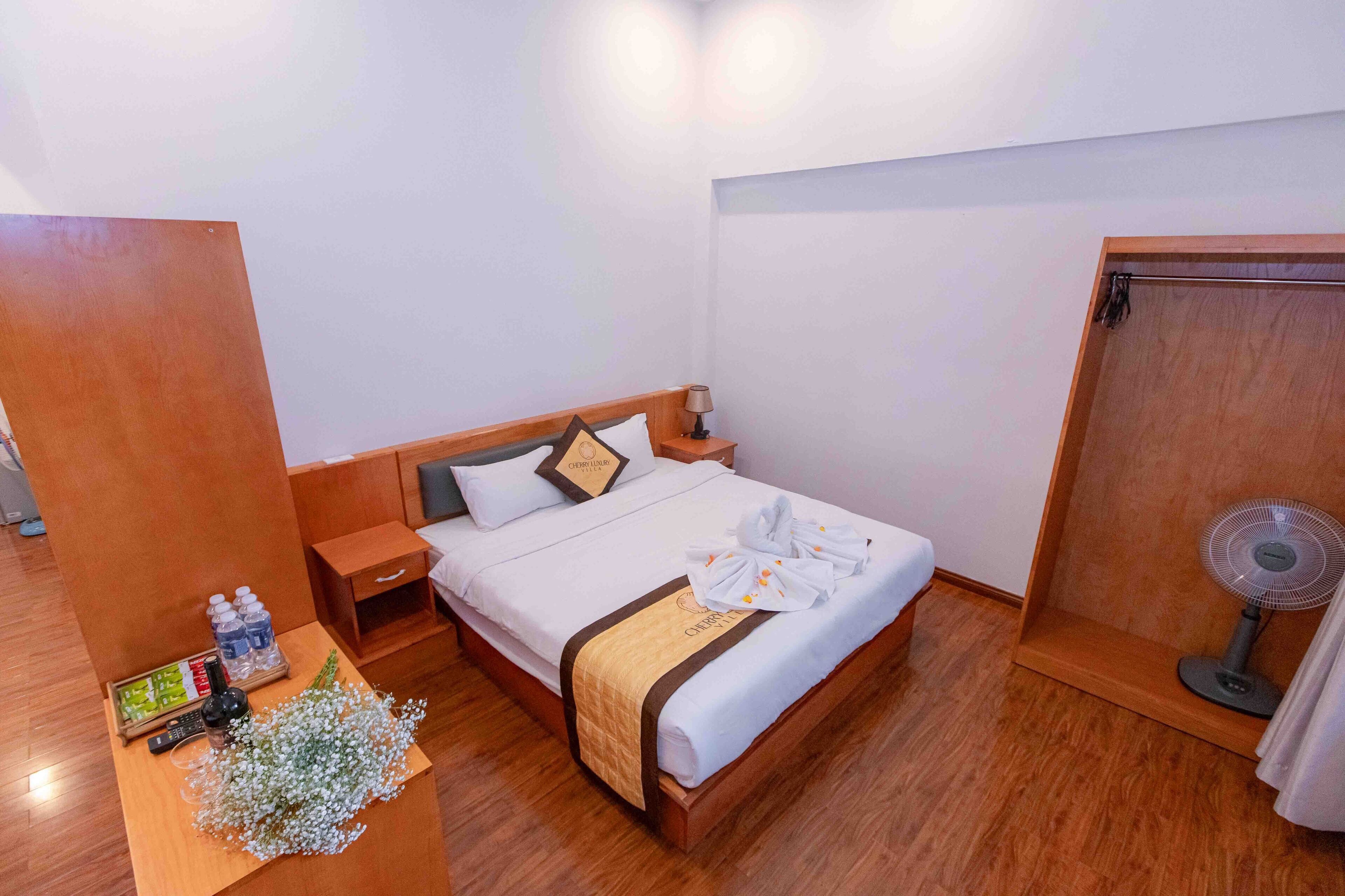 A neatly made bed with a wooden headboard, a bedside table with toiletries, a small desk with a remote control, and a wall-mounted fan.