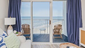 A room with a balcony overlooking the beach, furnished with a sofa, chairs, and a small table.