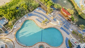An aerial view of a resort with a large central pool, multiple lounge areas with blue chairs, and a surrounding deck area.