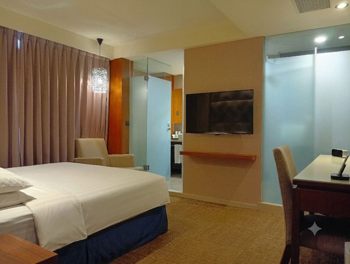 Habitación de hotel con cama, escritorio, silla, televisión y baño visible a través de un panel de vidrio.