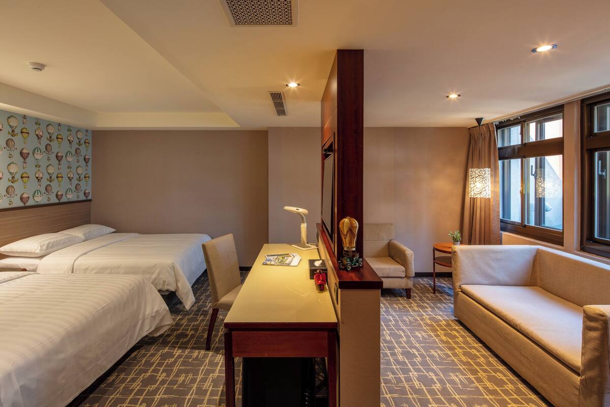 A hotel room with two beds, a desk, a sofa, and a large window.