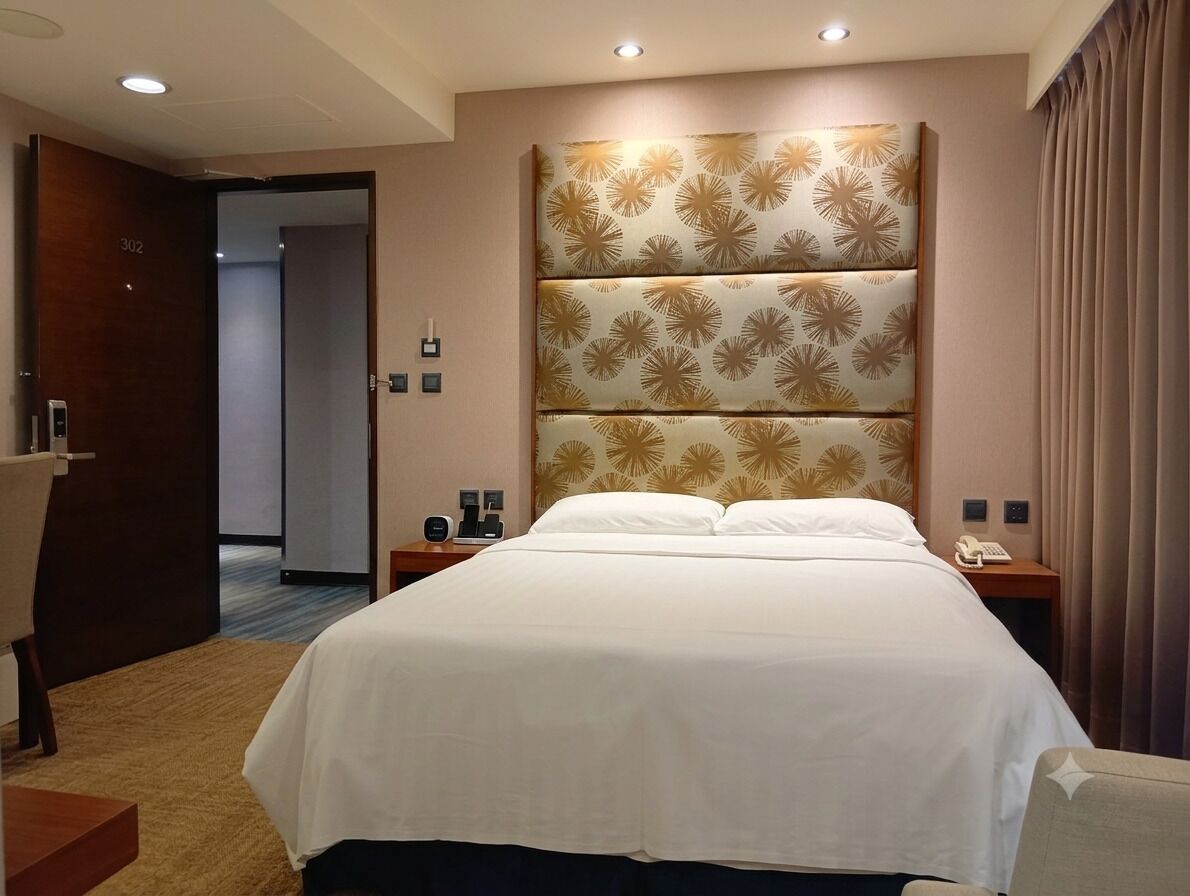 A hotel room with a large bed, a headboard with a floral pattern, bedside tables with lamps, and a door with a number.