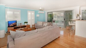 A modern living room with a beige sofa, a wooden coffee table, and a TV. There is a dining area with a white table and chairs, and a kitchen area with a white countertop and a phone. - Unit 4 'Marcoola Sands' on Petrie Avenue (Marcoola)