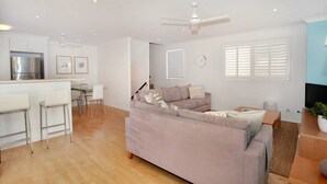A modern living room with a sofa, a dining area with a table and chairs, a kitchen with a refrigerator, and a ceiling fan. - Unit 4 'Marcoola Sands' on Petrie Avenue (Marcoola)