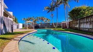 A swimming pool with a curved edge, surrounded by a wooden fence, palm trees, and a covered seating area. - Unit 4 'Marcoola Sands' on Petrie Avenue (Marcoola)