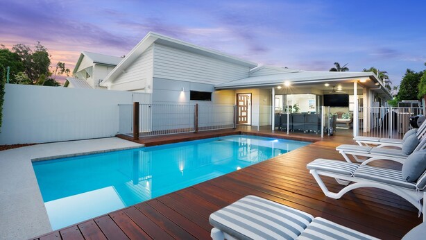 Ein Schwimmbecken mit Holzterrasse und Liegestühlen, umgeben von einem weißen Zaun und einem Haus mit überdachter Veranda.