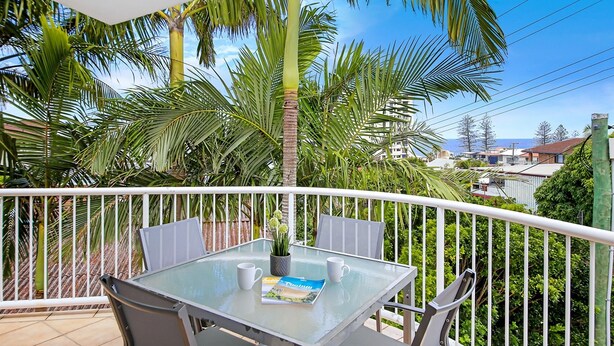 A glass-top table with two chairs, a potted plant, and a magazine on a balcony with white railings, surrounded by palm trees and a view of buildings. - Unit 8.12 Perry Street (Coolum Beach)