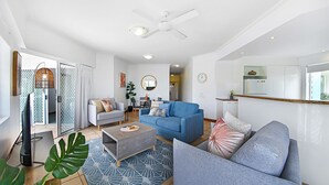 A modern living room with a grey sofa, a blue sofa, and a coffee table. There is a ceiling fan, a dining area in the background, and a kitchen with white cabinets. - Unit 8.12 Perry Street (Coolum Beach)