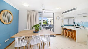 A modern kitchen with white cabinets, a central island with stools, and a dining area with a table and chairs. - Central Coolum Penthouse with Rooftop Terrace (Coolum Beach)