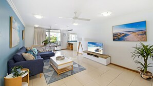 A modern living room with a sofa, coffee table, TV, and a dining area with chairs and a table. - Central Coolum Penthouse with Rooftop Terrace (Coolum Beach)