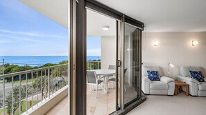 Un balcón con vista al océano, una mesa de comedor con sillas y dos sillones con una mesita y una lámpara.