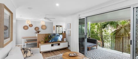A modern living room with a large TV, wooden furniture, and a balcony with a view of greenery.