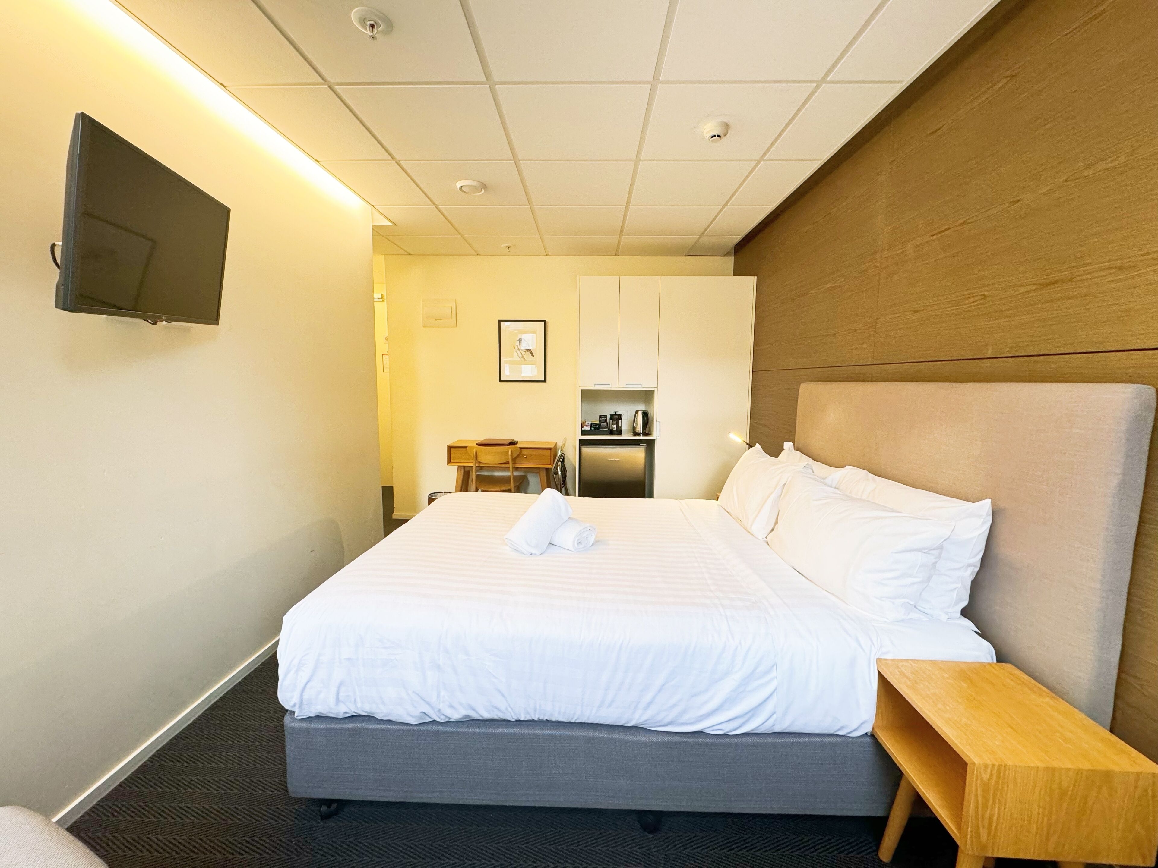 Une chambre d’hôtel dotée d’un grand lit, d’une table de chevet en bois, d’un bureau avec une chaise, d’un téléviseur fixé au mur et d’un coin cuisine.