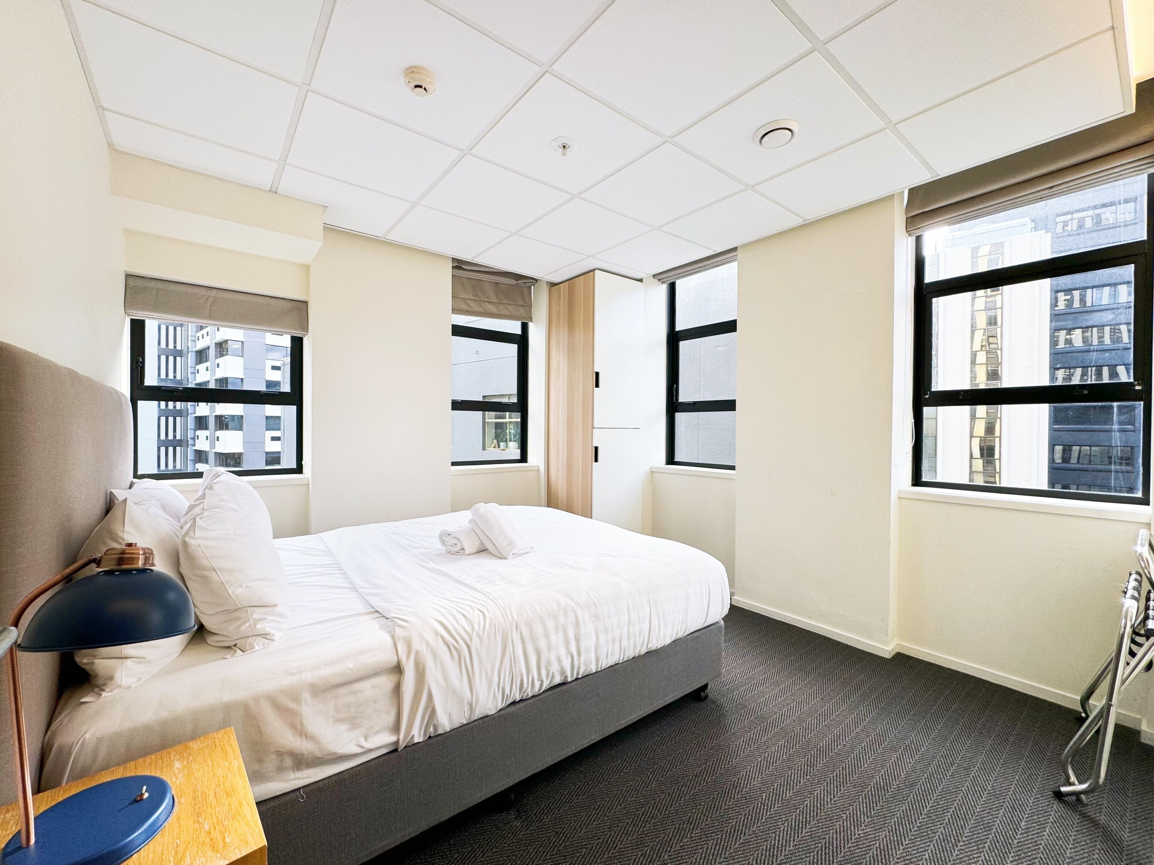 Een hotelkamer met een bed, een bureau met een lamp en grote ramen met uitzicht op de stad.