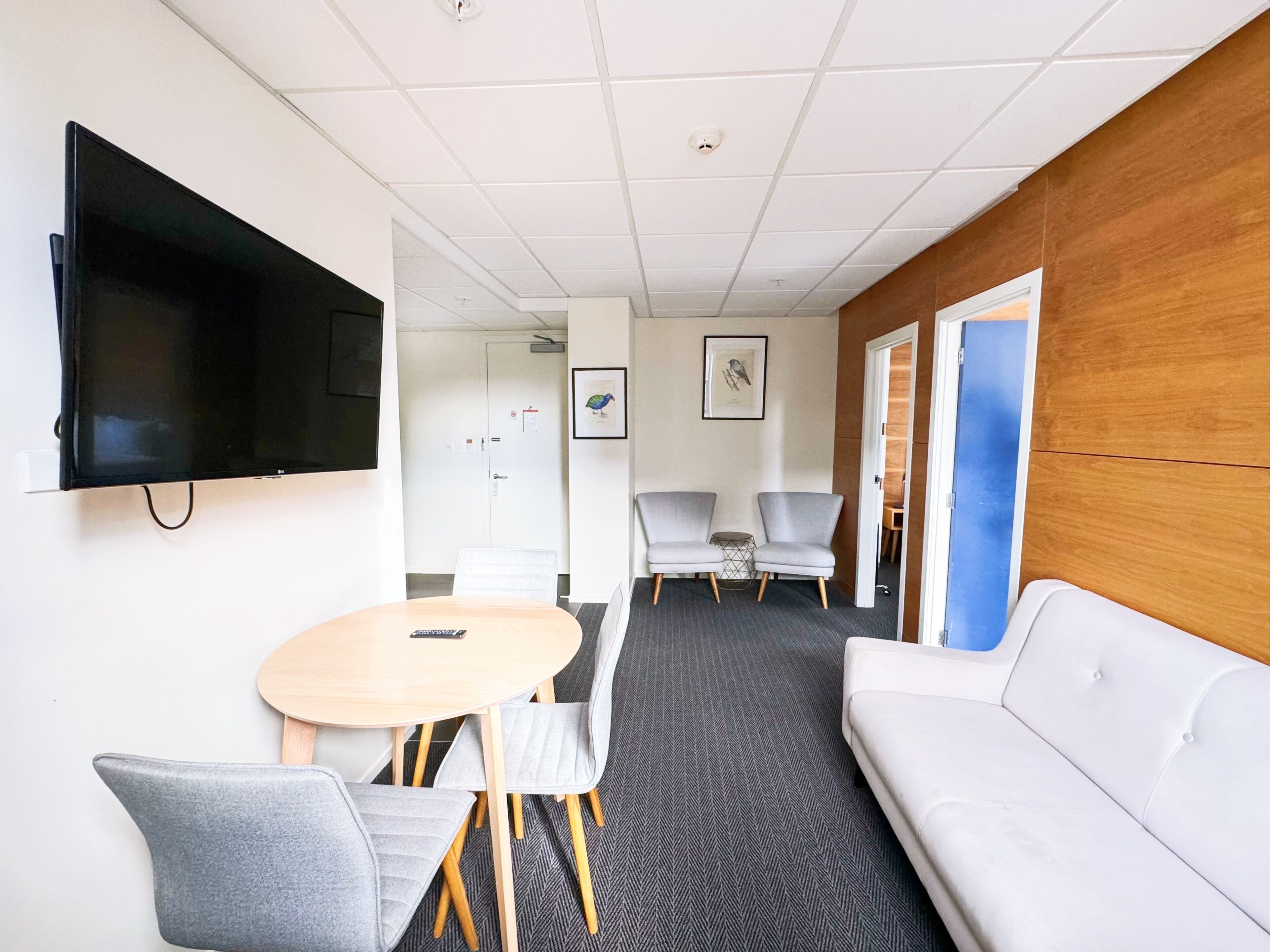 Ein modernes Hotelzimmer mit Flachbildfernseher, einem runden Tisch mit Stühlen, einer Couch und einer Wand mit Holzverkleidung.